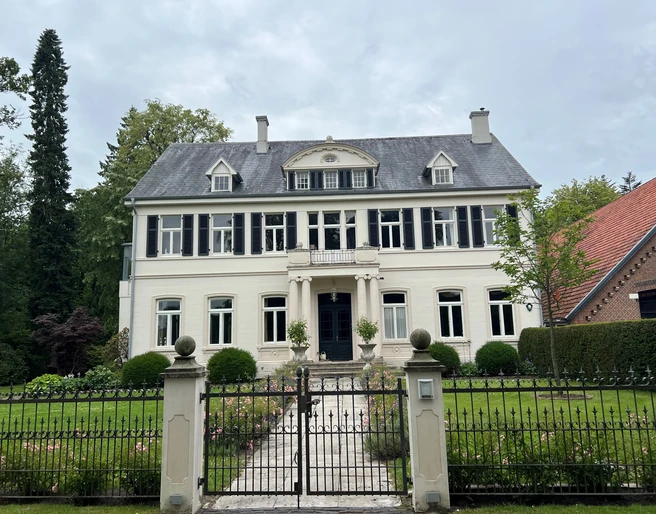 Bild5.jpg Ein großes, weißes Herrenhaus mit grauem Walmdach steht in einem gepflegten Garten hinter einem schmiedeeisernen Tor.