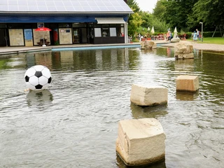 _MG_3583~2.JPG Ein Wasserbecken mit großen Steinen und einem riesigen aufblasbaren Fußball vor einem modernen Gebäude.