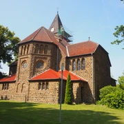 Eine historische Kirche aus Sandstein mit rotem Ziegeldach, umgeben von Grün, unter klarem Himmel.