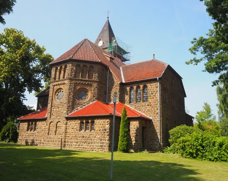 Eine historische Kirche aus Sandstein mit rotem Ziegeldach, umgeben von Grün, unter klarem Himmel.