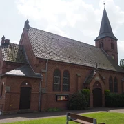 Die Christuskirche Oberbauerschaft, eine Backsteinkirche mit hohem spitzen Turm, steht sonnenbeschienen neben grünen Bäumen.