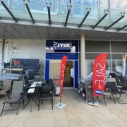 JYSK Stadtmitte, Wolfsburg.jpg Zu sehen ist die Eingangstür und die Schaufenster der Filiale JYSK. Die Schaufenster sind vollflächig mit Werbung beklebt. Vor dem Geschäft stehen verschiedene Gartenmöbel.