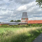 Wesermühle GbR Backsteinmühle mit zylinderförmigem Turm und roten Dächern vor bewölktem Himmel, umgeben von Wiesen.