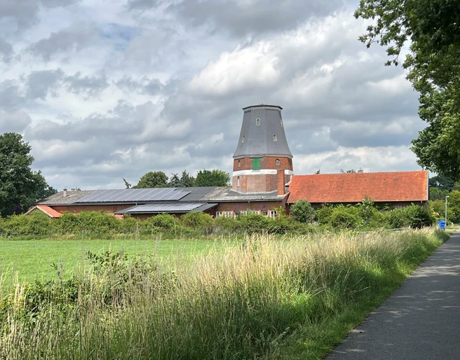 Wesermühle GbR Backsteinmühle mit zylinderförmigem Turm und roten Dächern vor bewölktem Himmel, umgeben von Wiesen.