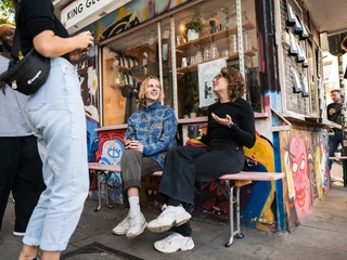 King Georg Kiosk Das Bild zeigt eine lebhafte Szene vor einem farbenfroh gestalteten Kiosk. Eine kleine Gruppe junger Menschen ist in entspannter Unterhaltung vertieft. Im Vordergrund sitzen zwei Personen auf einer Bank und genießen die lebendige Atmosphäre des Großstadtflairs.The picture shows a lively scene in front of a colorful kiosk. A small group of young people are engaged in relaxed conversation. In the foreground, two people sit on a bench and enjoy the lively atmosphere of the big city flair.