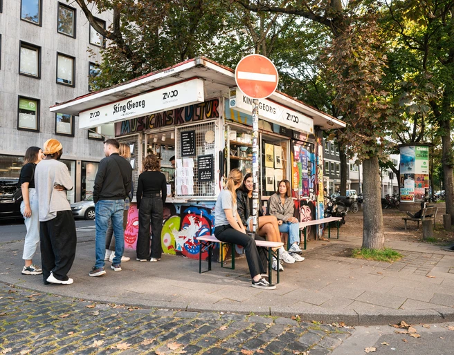 King Georg Kiosk Kleines Kiosk im urbanen Umfeld, umgeben von Bäumen und Menschen in entspannter Atmosphäre.Small kiosk in an urban setting, surrounded by trees and people in a relaxed atmosphere.