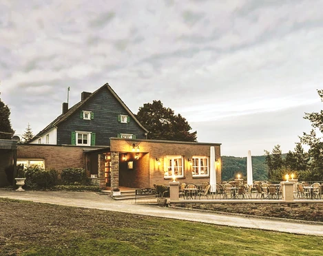 Restaurant Op dä Höh in Hattingen Ein Restaurant in einem Backsteingebäude mit Terrasse, Bäumen und bewölktem Himmel im Hintergrund.