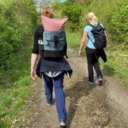 Zwei Frauen wandern auf einem schmalen Waldweg in sommerlichen Outfits und tragen Rucksäcke.