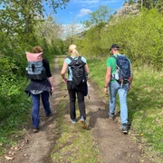 Drei Personen wandern auf einem naturbelassenen Weg, umgeben von frischem Grün und blauem Himmel.