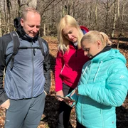 Drei Personen im Wald betrachten eine Karte bei sonnigem Wetter; alle tragen Outdoor-Kleidung.