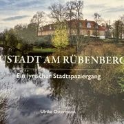 Geschichten aus Neustadt - Lesung mit Lyrik und Anekdoten Ein Buchcover zeigt einen ruhigen Fluss in Neustadt am Rübenberge, gesäumt von Bäumen und einem großen Backsteingebäude im Hintergrund.
