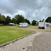 Wohnmobilstellplatz Heisterholz Grüne Wiese mit mehreren geparkten Wohnmobilen unter bewölktem Himmel, umgeben von Bäumen.
