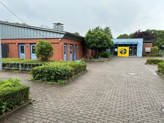 Schwimmbad mit blau-rotem Gebäude und gelber Eingangstür, umgeben von grünen Büschen und Bäumen.