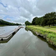 Schiffsanleger Heisterholz Schiffsanleger neben ruhigem Fluss, umgeben von grünen Wiesen und Bäumen unter bewölktem Himmel.