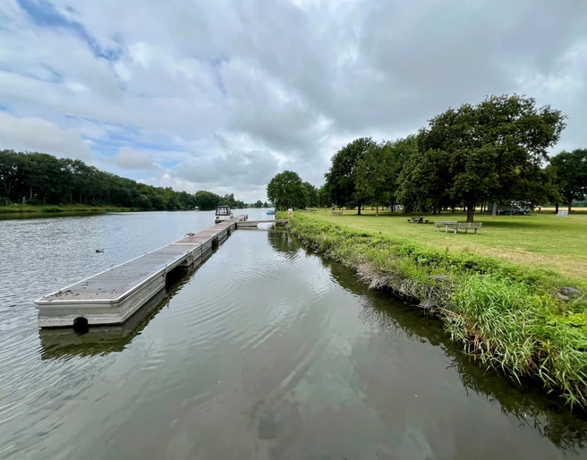 Schiffsanleger Heisterholz Schiffsanleger neben ruhigem Fluss, umgeben von grünen Wiesen und Bäumen unter bewölktem Himmel.