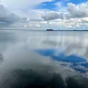 Weitblick auf ruhigen See mit spiegelnden Wolken und fernen kleinen Inseln unter wechselhaftem Himmel.