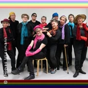 Eine fröhliche Gruppe älterer Menschen in bunter Kleidung, einige mit Hüten und Schals, posiert vor einer Wand. Sie tragen Accessoires in verschiedenen Farben, die Lebensfreude ausstrahlen.