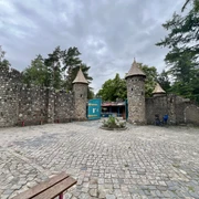 Magic Park Verden Ein steinernes Burgtor mit drei Türmen vor einem bewölkten Himmel, umgeben von Bäumen und Gehweg.