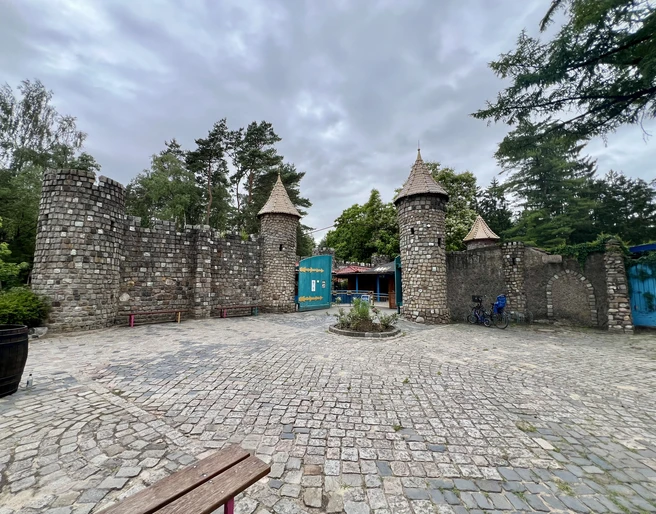 Magic Park Verden Ein steinernes Burgtor mit drei Türmen vor einem bewölkten Himmel, umgeben von Bäumen und Gehweg.