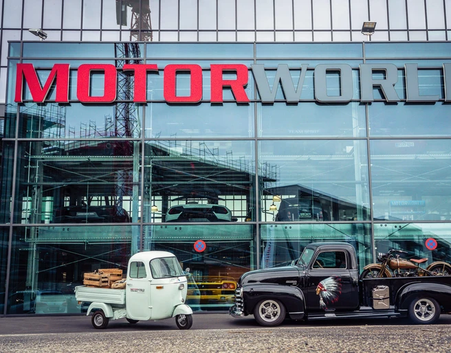 Motorworld Cologne Ein schwarzer Pickup-Truck mit einem Motorrad auf der Ladefläche und ein weißer Caddy stehen vor der gläsernen Fassade der Motorworld Köln, auf der das rot-graue Motorworld-Logo prangt.A black pickup truck with a motorcycle on the back and a white Caddy are parked in front of the glass façade of Motorworld Köln, emblazoned with the red and gray Motorworld logo.