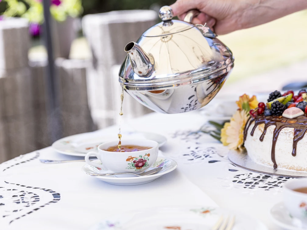 Teetied Gerade wird Tee aus einer eleganten Metallkanne in eine Porzellantasse gegossen, während eine üppige Sahnetorte mit frischen Früchten verziert danebensteht.