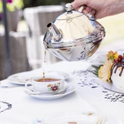 Teetied Gerade wird Tee aus einer eleganten Metallkanne in eine Porzellantasse gegossen, während eine üppige Sahnetorte mit frischen Früchten verziert danebensteht.