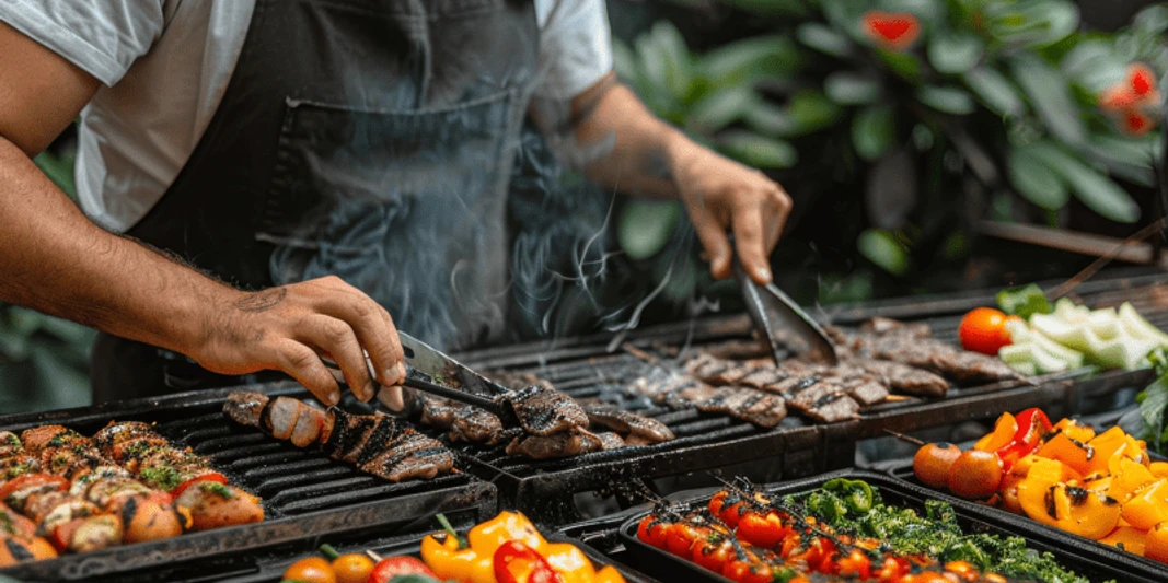 BBQ - Mit Live Cooking auf der Terrasse Grillmeister in schwarzer Schürze, der auf einem Barbecue-Grill verschiedene Fleischsorten und buntes Gemüse mit Grillzangen zubereitet, umgeben von frischen Zutaten und Rauchschwaden.Barbecue master in a black apron preparing various types of meat and colorful vegetables with barbecue tongs on a barbecue grill, surrounded by fresh ingredients and billows of smoke.