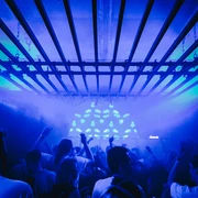 Bootshaus Dreherei Das Bild zeigt eine lebhafte Clubszene mit tanzenden Menschen unter blauer Beleuchtung und einem metallischen Gitter.The picture shows a lively club scene with people dancing under blue lighting and a metallic grid.
