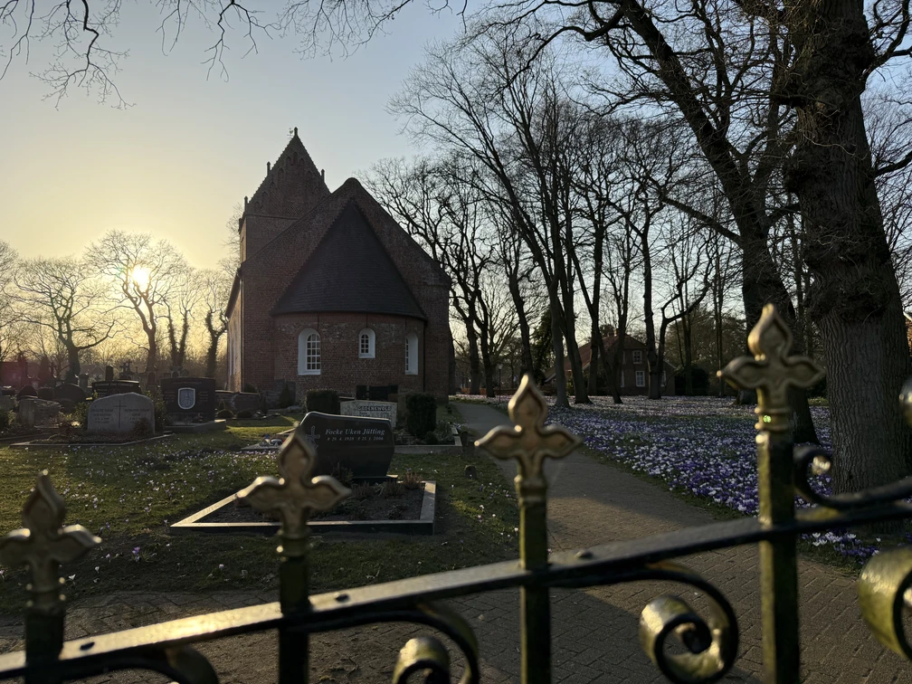 Kirche Backemoor Kirche mit Rundbogenfenstern und Friedhof im Sonnenuntergang, Bäume ohne Blätter und blühende Blumen am Weg