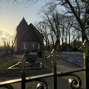 Kirche Backemoor  Kirche mit Rundbogenfenstern und Friedhof im Sonnenuntergang, Bäume ohne Blätter und blühende Blumen am Weg
