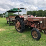 Hof Claus  Ein rustikaler roter Traktor mit Anhänger steht auf einer grünen Wiese unter wolkigem Himmel.
