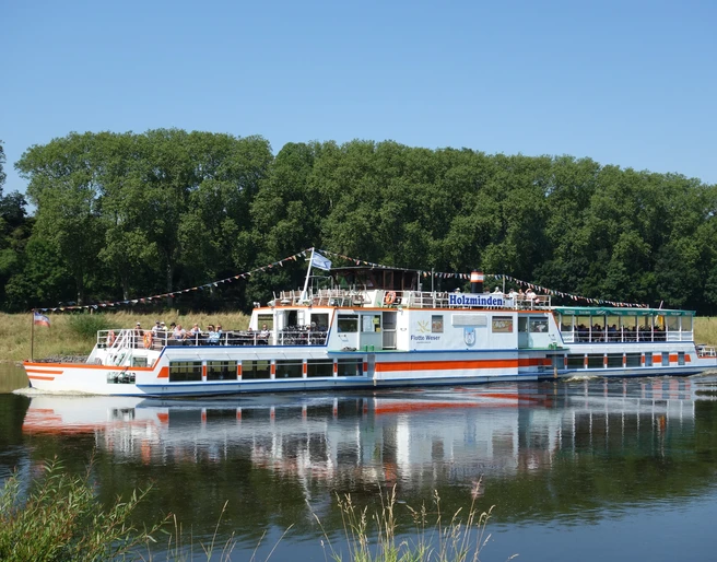 Flotte Weser Das Ausflugsschiff Flotte Weser fährt auf einem Fluss, gesäumt von grünen Bäumen.