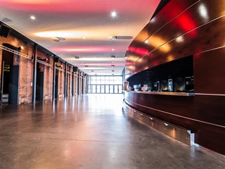 Palladium Köln Foyer Das Bild zeigt das Foyer des Palladium Köln. Der Raum hat eine geschwungene Holztheke rechts und offen gelegte Backsteinwände links. Die Decke ist mit einer modernen Lichtinstallation beleuchtet, die warme, rötliche Töne widergibt. Der Boden besteht aus glattem Beton, was eine moderne, industrielle Atmosphäre schafft. Im Hintergrund befindet sich ein großer Fensterbereich, durch den Tageslicht einfällt und den Raum erhellt.The picture shows the foyer of the Palladium Cologne. The room has a curved wooden counter on the right and exposed brick walls on the left. The ceiling is illuminated with a modern light installation that reflects warm, reddish tones. The floor is made of smooth concrete, creating a modern, industrial atmosphere. In the background is a large window area, allowing daylight to flood in and brighten the room.