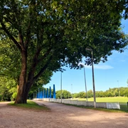 Turnierplatz Tribuenen.jpg Das Bild zeigt eine breite, von Bäumen gesäumte Allee neben einem offenen Sportplatz mit blau-weißen Tribünen.