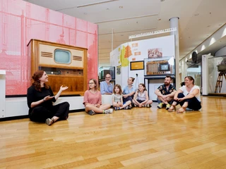 Group tour at the Museum of Communication Frankfurt in front of a historic television Eine Gruppe von Besucher*innen sitzt auf dem Boden vor einem historischen Fernseher und hört aufmerksam einer Führerin zu.A group of visitors sitting on the floor in front of a historic TV, attentively listening to a guide.