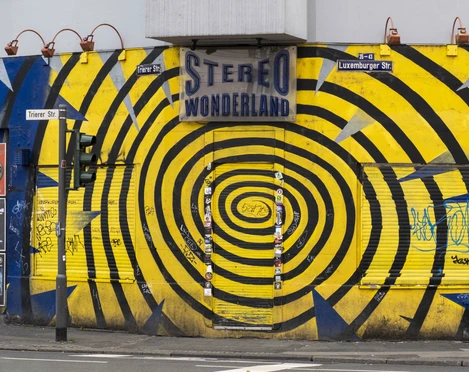 Stereo Wonderland Das Bild zeigt die Vorderseite eines Gebäudes mit einem markanten gelben und blauen Spiralmuster. An der Fassade befindet sich ein Schild mit der Aufschrift "Stereo Wonderland". Graffiti und Plakate sind auf dem Rolltor und den Wänden zu sehen, was eine urbane und künstlerische Atmosphäre erzeugt.The picture shows the front of a building with a striking yellow and blue spiral pattern. There is a sign on the façade that reads "Stereo Wonderland". Graffiti and posters can be seen on the roller shutter and the walls, creating an urban and artistic atmosphere.