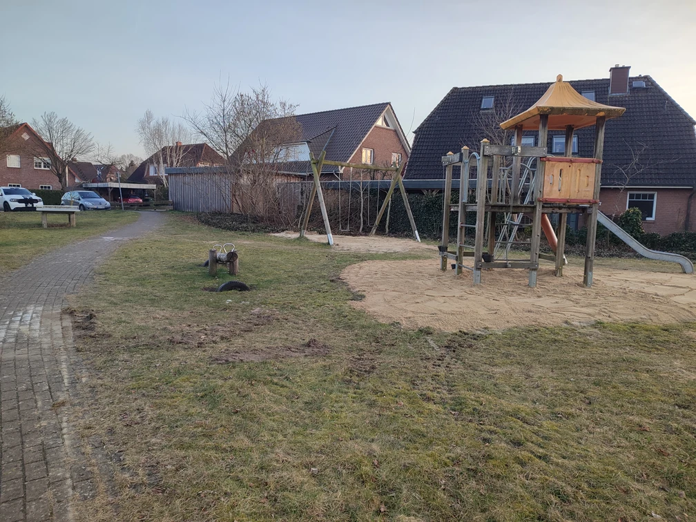 Spielplatz an den Weiden Ein Spielplatz An den Weiden mit Holz-Klettergerüst und Rutsche, umgeben von Gras und einigen Häusern im Hintergrund.