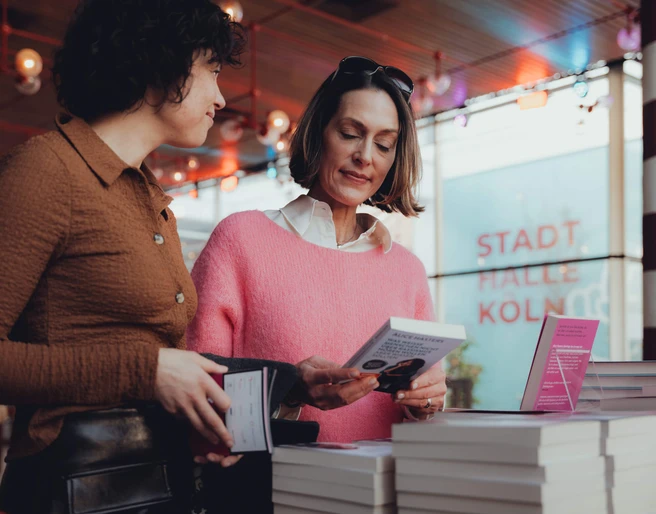 lit.COLOGNE Zwei Frauen studieren Bücher in einem modernen Ausstellungsraum. Im Hintergrund leuchtet "Stadthalle Köln".Two women study books in a modern exhibition space. "Stadthalle Köln" shines in the background.