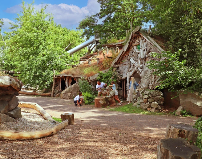 ArkaZien Erlebniswelt Region Leipzig - Bergwerk Kinder erforschen einen grasüberwachsenen Eingang eines verlassenen Holzbergwerks, umgeben von Bäumen.