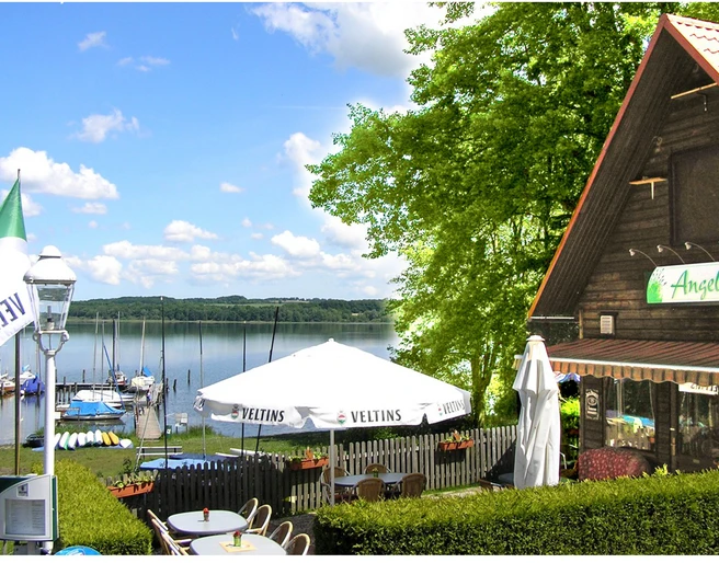 Naturcamping Buchholz_Mühle Ein Campingplatz am See mit grünem Ufer, Segelbooten und einem holzverkleideten Restaurant im Vordergrund.