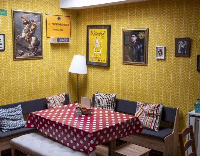 The Bär Das Bild zeigt einen Raum mit gelben Tapeten, einer Sitzecke mit Polsterkissen und dekorativen Bildern an der Wand. Ein Tisch mit rotem Punktetuch steht zentral, ergänzt durch bequeme Holzstühle.The picture shows a room with yellow wallpaper, a seating area with upholstered cushions and decorative pictures on the wall. A table with a red polka dot cloth stands in the center, complemented by comfortable wooden chairs.