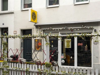 The Bär Ein Café in Köln mit weißer Fassade und einladendem Außenbereich. Bunte Stühle stehen vor einem weißen Zaun, der mit grünen Pflanzen dekoriert ist.A café in Cologne with a white façade and inviting outdoor area. Colorful chairs stand in front of a white fence decorated with green plants.