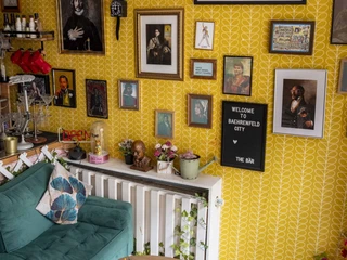 The Bär In einem Raum mit gelber Mustertapete hängen zahlreiche Kunstwerke. Ein Sofa in Petrolfarbe, verziert mit stilvollen Kissen, steht vor einem schmalen Wandtisch. Auf diesem finden sich Pflanzen und Skurrilitäten. Ein kleiner Holztisch mit Büchern und Magazinen ergänzt das künstlerische Ensemble.Numerous works of art hang in a room with yellow patterned wallpaper. A petrol-colored sofa, adorned with stylish cushions, stands in front of a narrow wall table. Plants and curiosities are placed on it. A small wooden table with books and magazines completes the artistic ensemble.