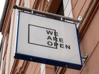 TRINK-GENOSSIN Ein rechteckiges Schild mit der Aufschrift „WE ARE OPEN“ hängt an einer rosafarbenen Hausfassade.A rectangular sign with the inscription "WE ARE OPEN" hangs on a pink-colored house facade.