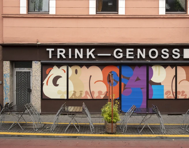 TRINK-GENOSSIN Fassade eines Kölner Lokals mit buntem Wandkunstwerk und leeren Tischen im Vordergrund.Facade of a Cologne restaurant with colorful wall art and empty tables in the foreground.