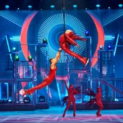Akrobatische Eiskunstläuferinnen und -läufer in glitzernden roten Kostümen schweben elegant durch die Luft auf einer farbenfrohen Bühne mit neonbeleuchteten Hintergründen.Acrobatic figure skaters in glittering red costumes float elegantly through the air on a colorful stage with neon-lit backdrops.