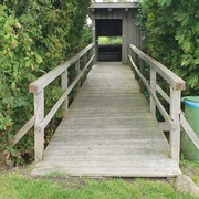 Vogelbeobachtungshütte  Eine Vogelbeobachtungshütte aus Holz mit überdachtem Zugang und umgeben von grünen Büschen.