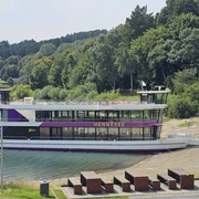 EMS Hennesee Ein weißes Ausflugsschiff mit zwei Decks liegt an einem bewaldeten Ufer des Hennesees an.