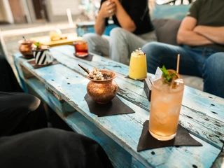 Toddy Tapper Das Bild zeigt eine Gruppe von Menschen, die draußen an einem Tisch aus blauen Holzbrettern sitzen. Vor ihnen stehen dekorative Getränke in verschiedenen Gläsern. Es herrscht eine entspannte Atmosphäre.The picture shows a group of people sitting outside at a table made of blue wooden boards. In front of them are decorative drinks in various glasses. The atmosphere is relaxed.