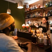Toddy Tapper Eine Barkeeperin mixt Cocktails in einer atmosphärischen Bar vor aufmerksamem Publikum.A bartender mixes cocktails in an atmospheric bar in front of an attentive audience.